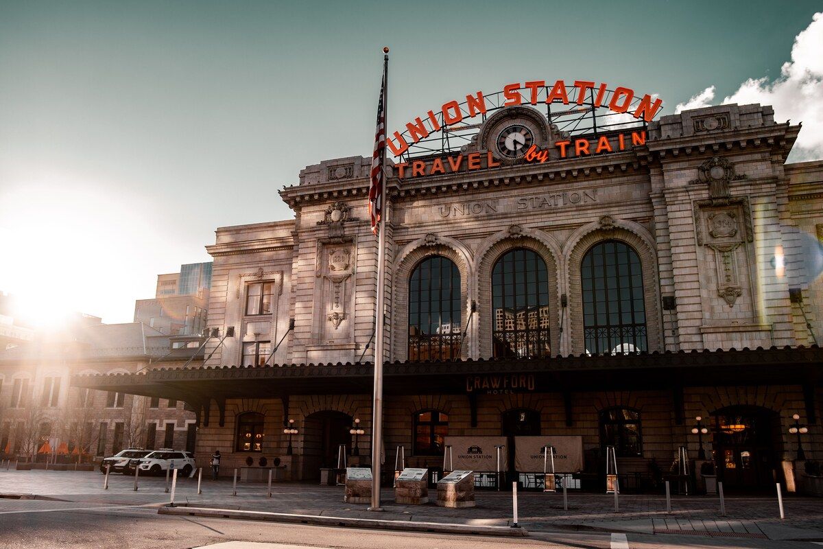Denver Union Station from the street, its arched neon roof sign lettered UNION STATION and TRAVEL BY TRAIN above a clock.
