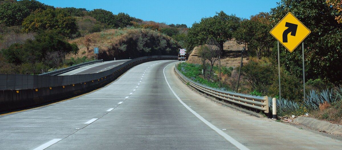 A divided highway curving through dry green hills past a yellow curve warning sign, a road a Puerto Vallarta transfer drives.